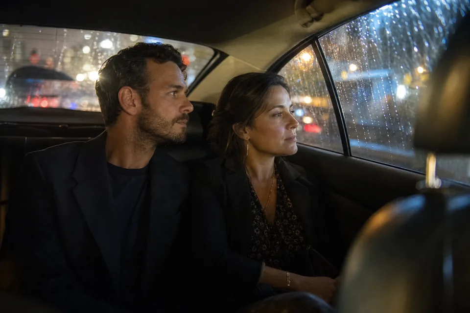 Adult couple close together in the back seat of a taxi at night, city lights blurred through the wet window