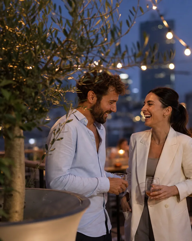 Man and woman in their thirties talking on a rooftop terrace at dusk with drinks, relaxed and flirty